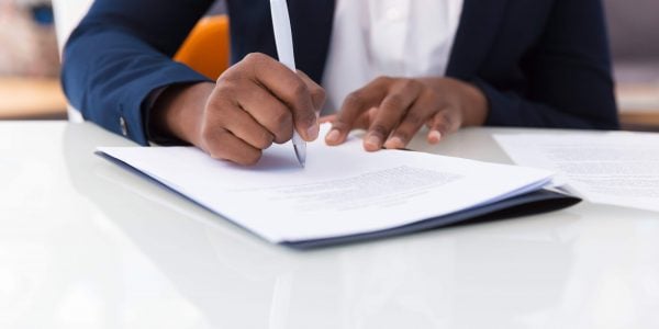Businesswoman signing contract. African American business woman sitting at table in office, holding pen and writing in document. Legal expertise concept