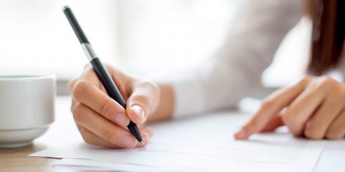 Hand of young businesswoman writing on paper or signing contract at table in office